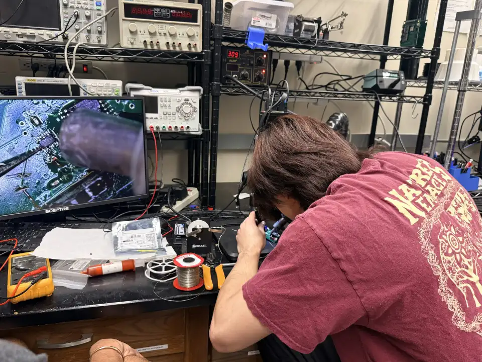 A team member, seen from behind, bends over an inspection microscope at a repair bench; the board appears magnified on a monitor beside a multimeter, solder and tweezers.